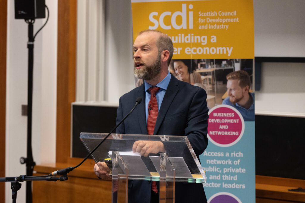 Jonathan speaking at a clear podium. In the background is a yellow-headed banner with the SCDI's logo.